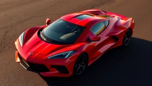 Red 2024 Chevrolet Corvette C8 mid-engine sports car photographed at three-quarter front angle on dark asphalt background, sunlight reflecting off aerodynamic body panels and distinctive split rear window design